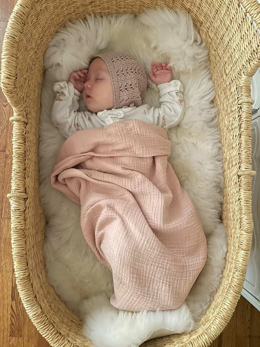 Newborn baby wrapped in a pink blanket and wearing a knitted hat, lying in a bright ivory nursery sheepskin