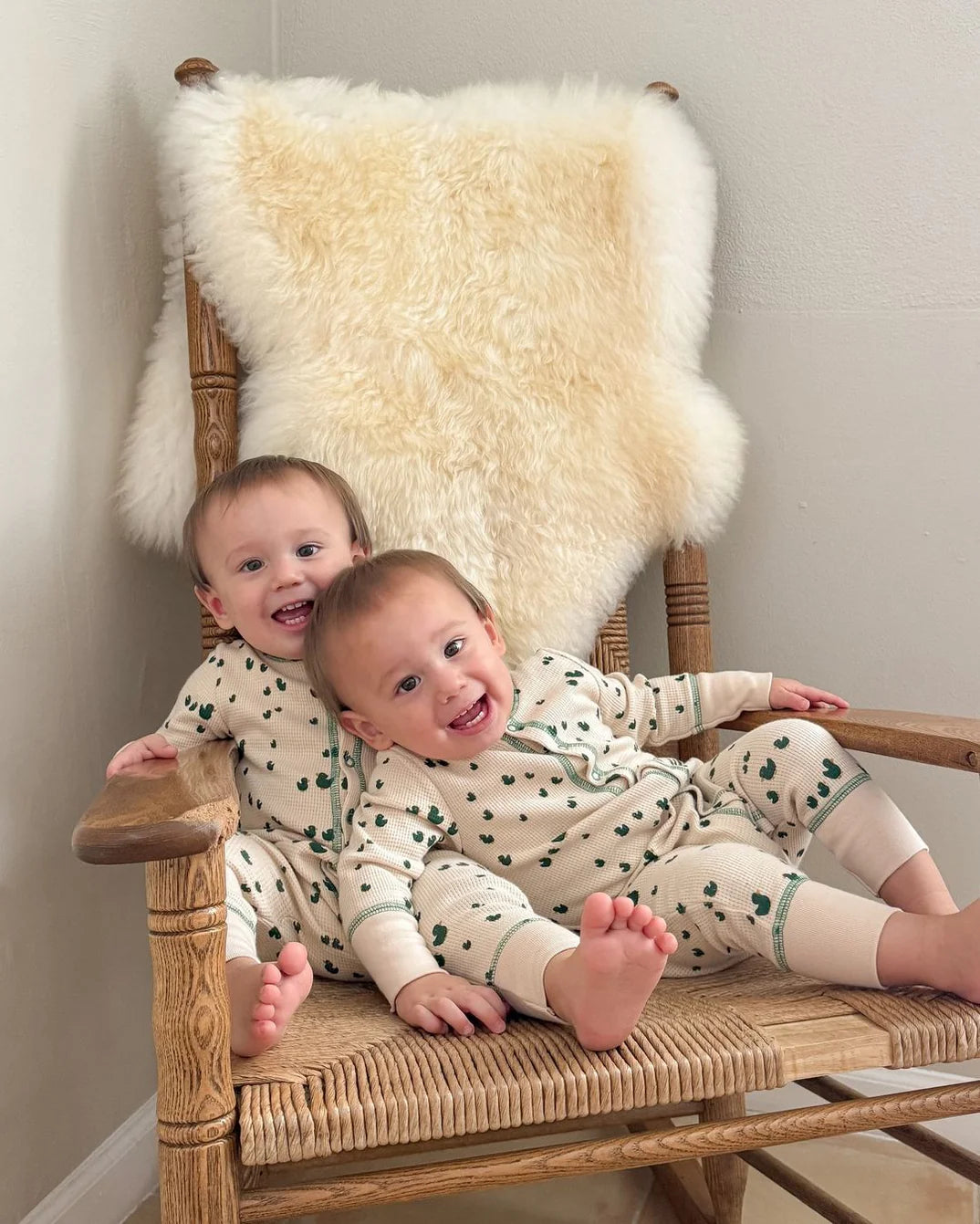 Two children in matching outfits sitting on a wooden chair with a bright ivory nursery sheepskin