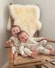 Two children in matching outfits sitting on a wooden chair with a bright ivory nursery sheepskin