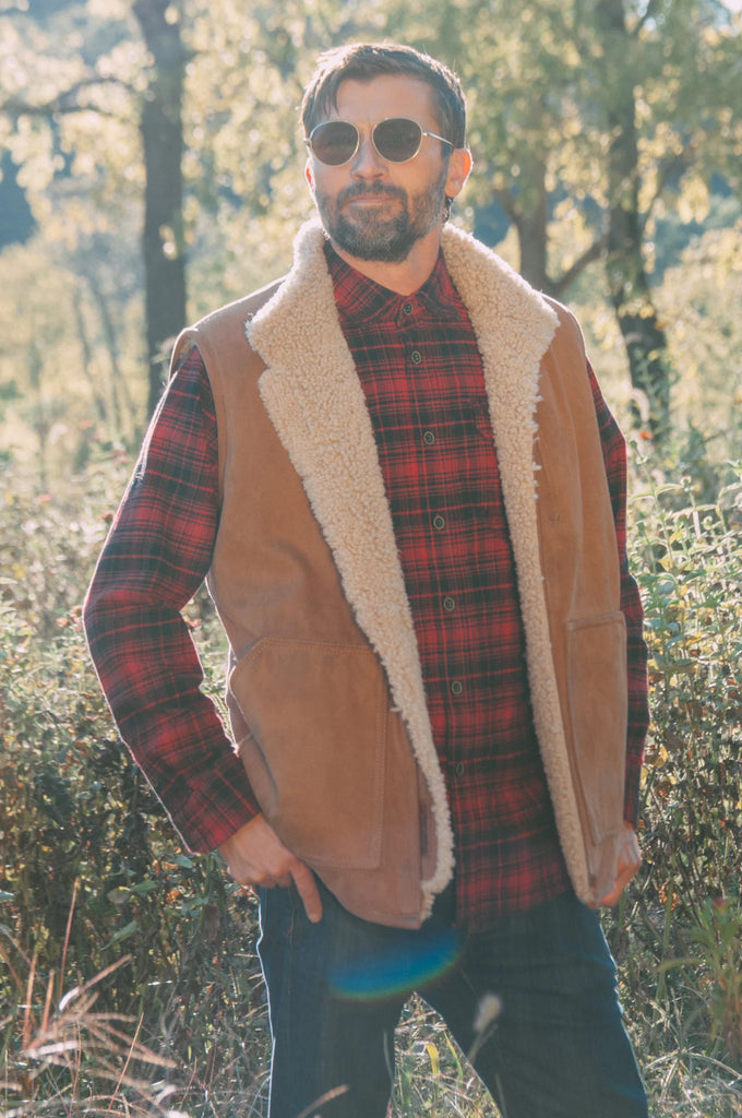 Man wearing a brown sheepskin mens vest over a plaid shirt in a forest setting