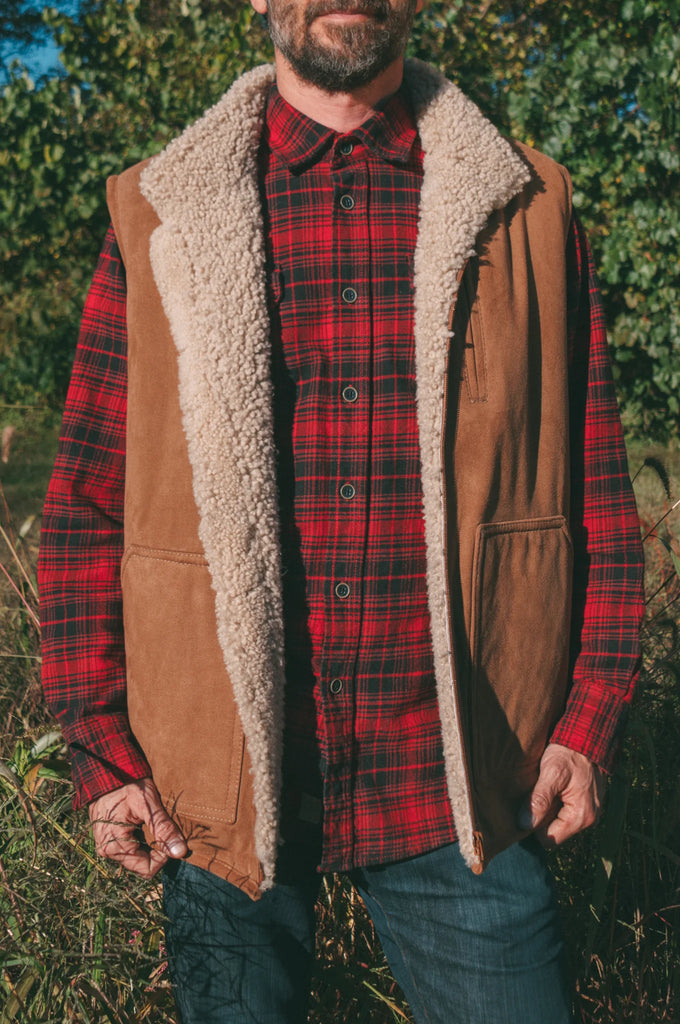 Person wearing a red plaid shirt, brown sheepskin mens vest with white fur lining, and blue jeans outdoors.